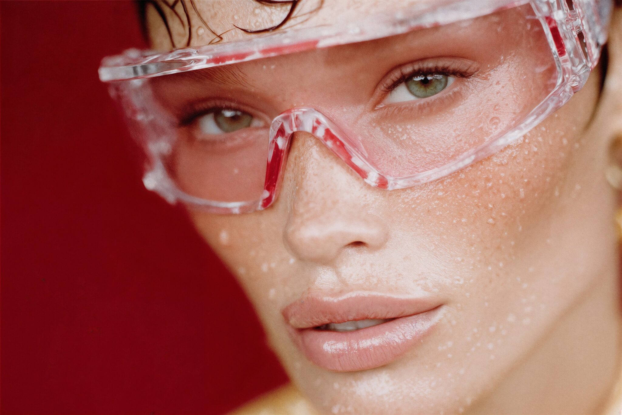AI beauty visual of model with wet skin and translucent glasses on red background, photoreal luxury editorial tone