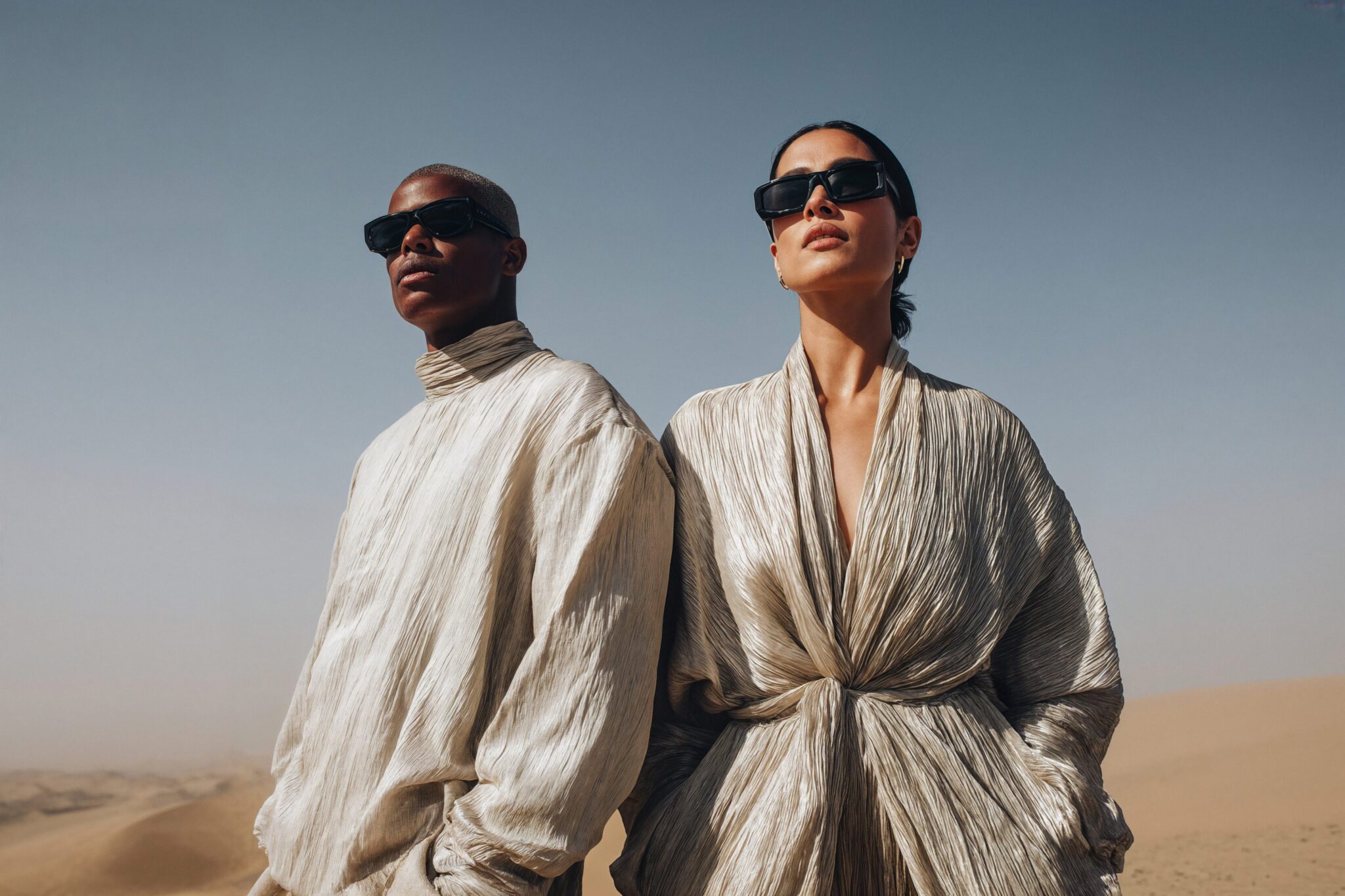 AI fashion visual of two models wearing pleated metallic outfits in desert light, cinematic campaign tone